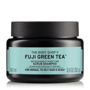 A short cylindrical black plastic Tub with a black plastic llid and a dark green label that has The Body Shop Fuji Green Tea™ Scrub Shampoo written in bright green writing and some pictures of some bright green tea leaves on it, on a white background.