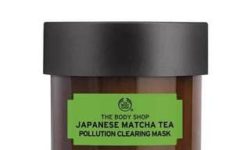 A short cylindrical clear glass Jar full of some creamy green liquid with a matte black plastic lid and a green label that has The Body Shop Japanese Matcha Tea Pollution Clearing Mask written in bold black writing on it, on a white background.