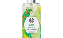 A tall cylindrical clear plastic bottle full of some clear liquid with a metallic silver spray lid and a clear label that has The Body Shop Lime & Matcha Hair & Body Mist written in bold black writing and some pictures of some bright green limes on it, on a white background.