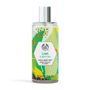 A tall cylindrical clear plastic bottle full of some clear liquid with a metallic silver spray lid and a clear label that has The Body Shop Lime & Matcha Hair & Body Mist written in bold black writing and some pictures of some bright green limes on it, on a white background.