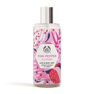 A tall cylindrical clear plastic bottle full of some clear liquid with a metallic silver spray lid and a clear label that has The Body Shop Pink Pepper & Lychee Hair & Body Mist written in bold black writing and some pictures of some bright pink lychees on it, on a white background.