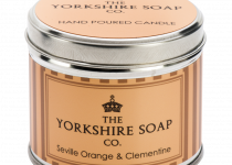 A short cylindrical silver tin with a silver lid and a bright orange label that has The Yorkshire Soap Company Seville Orange & Clementine Tin Candle written in bold black writing and a picture of a black crown on it, on a white background.