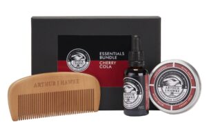 A large rectangular black cardboard box that has Arthur J Hawke Cherry Cola Essentials written in bold white writing on it next to a large dark brown semicircular beard comb , a short rectangular brown glass bottle with a black rubber pipette lid and a black label that has Arthur J Hawke Cherry Cola Beard Oil written in bold white writing on it and a short cylindrical metal tin with a black label that has Arthur J Hawke cherry cola beard balm written in bold white writing on it, on a white background.