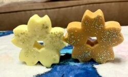 A large bright orange flower shaped wax melt that's covered in some silver lustre next to a large neon yellow flower shaped wax melt that's covered in some silver lustre, on a white background.