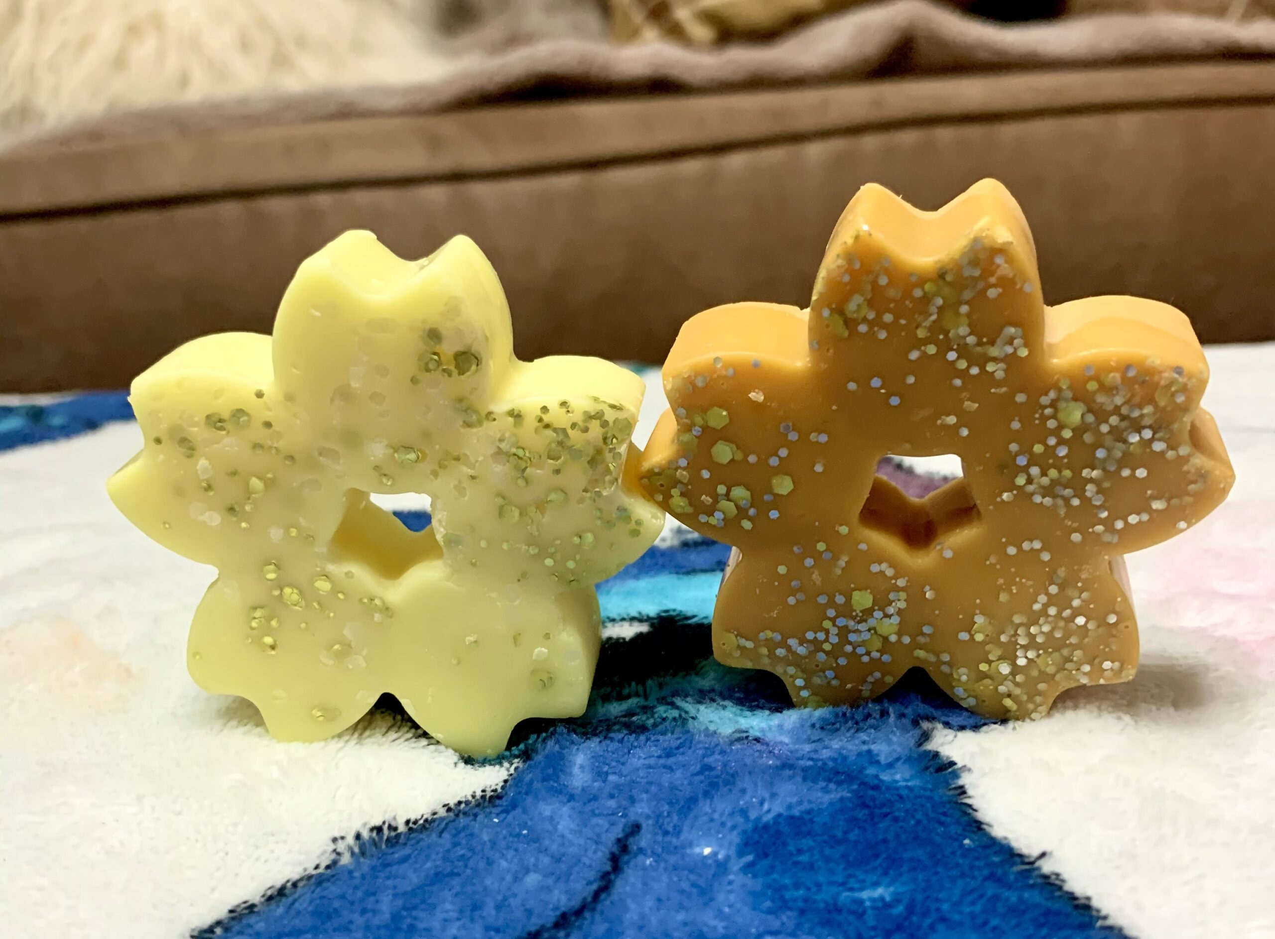 A large bright orange flower shaped wax melt that's covered in some silver lustre next to a large neon yellow flower shaped wax melt that's covered in some silver lustre, on a white background.