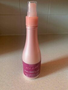 A tall cylindrical dark pink Plastic Wine Bottle with a dark red label that has Hask Red Wine 10 In 1 Leave In Conditioning Spray written in bold white writing on it, on a white background.