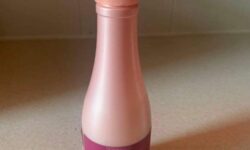A tall cylindrical dark pink Plastic Wine Bottle with a dark red label that has Hask Red Wine 10 In 1 Leave In Conditioning Spray written in bold white writing on it, on a white background.