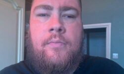 A tall bearded man that has some bright orange liquid all over his face wearing a light grey v-neck t-shirt stood against a white door.