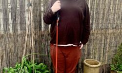 A tall bearded man wearing a chocolate brown roll neck top, a pair of dark brown chinos and a pair of shiny bright red shoes holding a bright red cane stood against a light brown brick wall and a dark brown wooden fence.