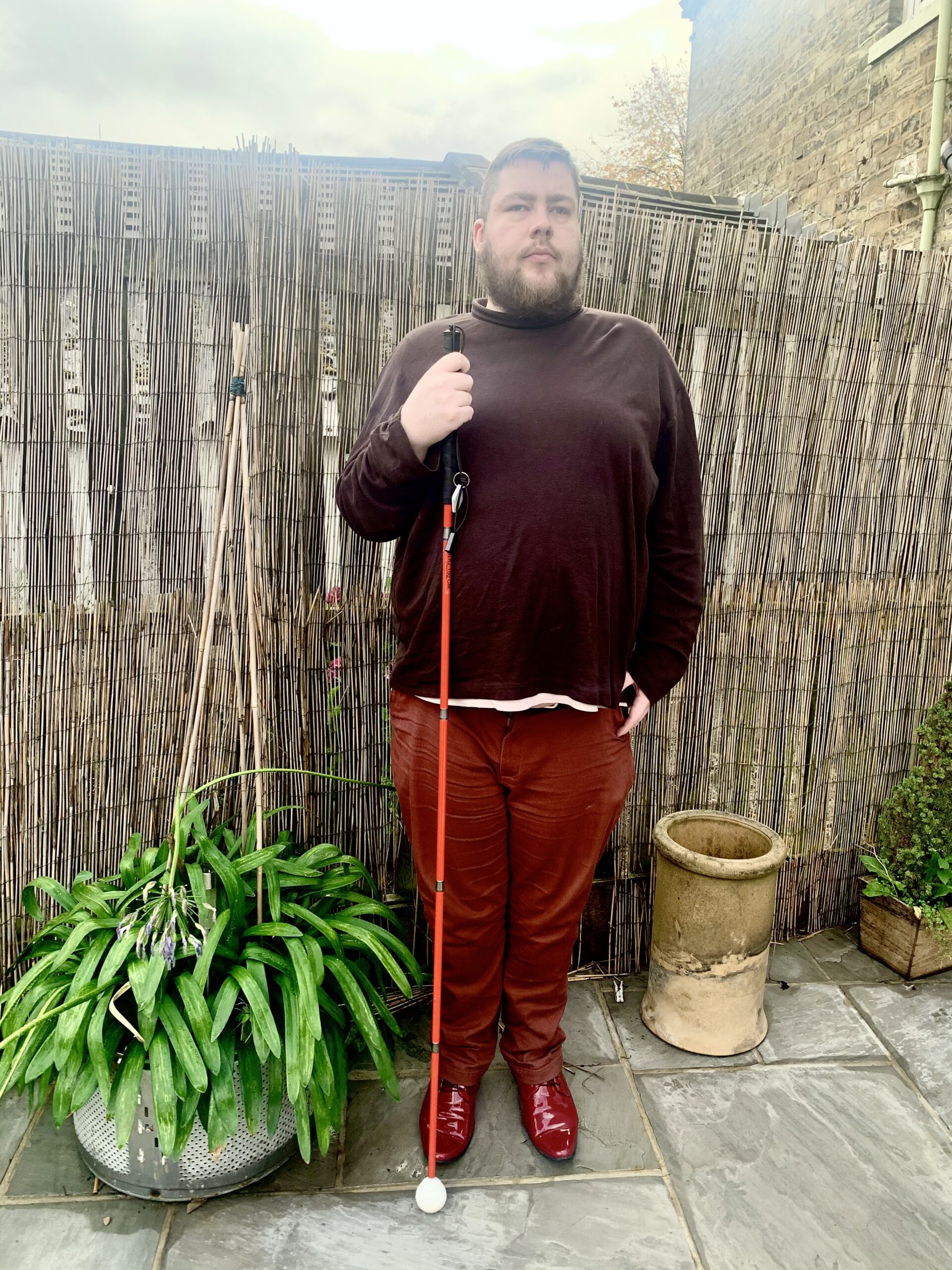 Outfit Of The Day | Casual Christmas Shopping A tall bearded man wearing a chocolate brown roll neck top, a pair of dark brown chinos and a pair of shiny bright red shoes holding a bright red cane stood against a light brown brick wall and a dark brown wooden fence.