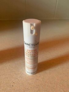 A tall cylindrical white plastic bottle that has Sanctuary Spa written in bold black writing and Power Peptide Awakening Eye Serum written in bright orange writing on it, on a white background.