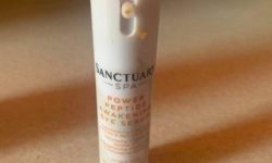 A tall cylindrical white plastic bottle that has Sanctuary Spa written in bold black writing and Power Peptide Awakening Eye Serum written in bright orange writing on it, on a white background.