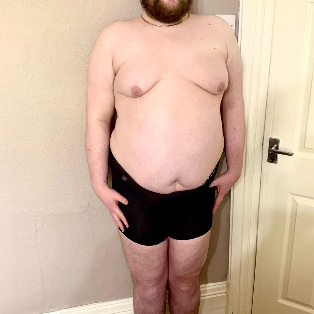 A tall bearded man wearing a pair of black trunks that has Step One written in bold white writing on them stood against a white door.