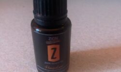 A short cylindrical dark brown glass bottle with a a Black Plastic Lid and a black label that has Zion Essentials Promised Land Beard Oil written in bold white writing and a picture of a bright orange stripe on it, on a white background.