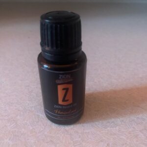 A short cylindrical dark brown glass bottle with a a Black Plastic Lid and a black label that has Zion Essentials Promised Land Beard Oil written in bold white writing and a picture of a bright orange stripe on it, on a white background.