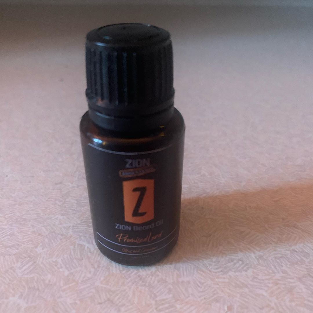 My Beard Has Reached The Promised Land | Zion Essentials Promised Land Beard Oil | Gifted A short cylindrical dark brown glass bottle with a a Black Plastic Lid and a black label that has Zion Essentials Promised Land Beard Oil written in bold white writing and a picture of a bright orange stripe on it, on a white background.