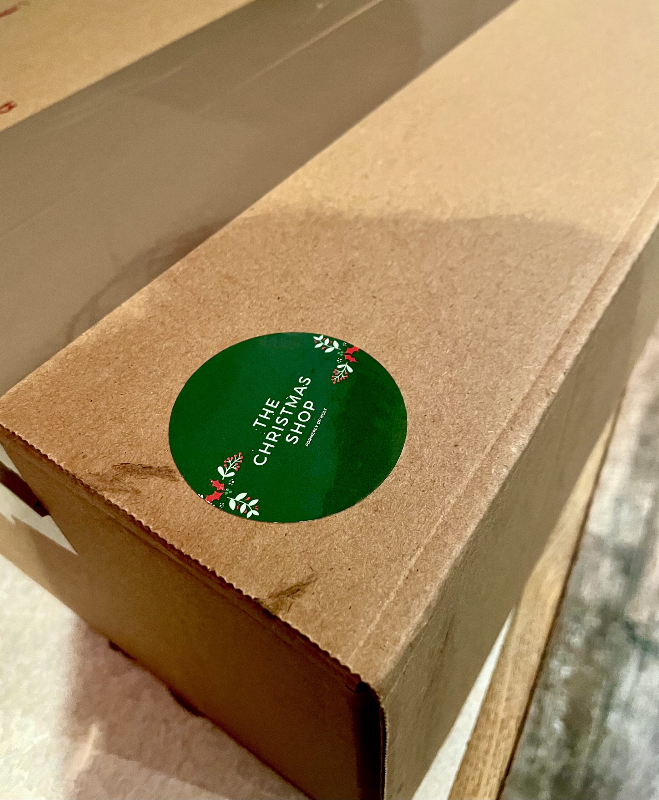 A large rectangular dark brown cardboard box with a bright green sticker that has Christmas Shop Holt written in bold white writing on it, on a white background.
