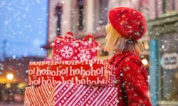 A tall woman with long blonde hair wearing a bright red hat and coat holding several rectangular red and white parcels walking in the city whilst it's snowing.