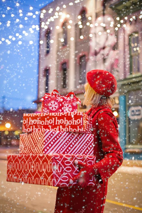 Luke Loves Christmas 2020 | Christmas 2020 Bloggers Collaborative Giveaway A tall woman with long blonde hair wearing a bright red hat and coat holding several rectangular red and white parcels walking in the city whilst it's snowing.