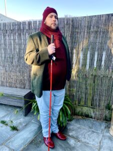 A tall bearded man wearing a dark red Jumper, a dark green tweed jacket, a dark red Beanie, a dark red scarf, a pair of stone grey chinos and a pair of shiny red shoes holding a bright red cane stood against a light brick wall.