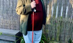 A tall bearded man wearing a dark red Jumper, a dark green tweed jacket, a dark red Beanie, a dark red scarf, a pair of stone grey chinos and a pair of shiny red shoes holding a bright red cane stood against a light brick wall.