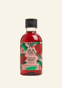 A tall cylindrical clear plastic bottle full of some florescent red liquid with a dark green lid and a white label that has The Body Shop written in bold black writing, Festive Berry Shower Gel written in bold dark red writing, and some pictures of some dark red berries on it, on a white background.