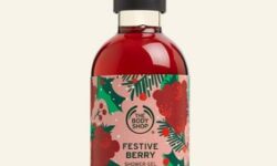 A tall cylindrical clear plastic bottle full of some florescent red liquid with a dark green lid and a white label that has The Body Shop written in bold black writing, Festive Berry Shower Gel written in bold dark red writing, and some pictures of some dark red berries on it, on a white background.