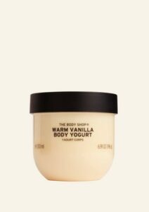 A short rounded rectangular clear plastic tub full of some creamy white liquid with a dark green lid that has The Body Shop Warm Vanilla Body Yoghurt written in dark green writing and some pictures of some dark brown vanilla pods on it, on a white background.