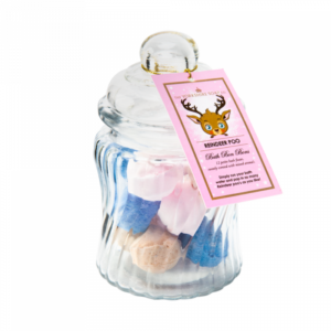 A small cylindrical clear glass jar with a clear glass lid and a white label that has The Yorkshire Soap Company Reindeer Poo Bath Bon Bons written in bold black writing and a picture of a large dark brown animated reindeers face on it, on a white background.