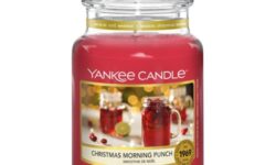 A tall cylindrical jar full of some dark red wax with a label that has Yankee Candle written in white writing, Christmas morning punch written in black writing, and a picture of a glass tumbler full of some dark red liquid surrounded by some out of focus fairy lights on it, on a white background.