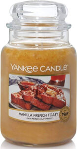 A tall cylindrical Jar full of some sandy yellow wax with a label that has Yankee Candle written in white writing, Vanilla French Toast written in black writing, and a picture of a large slice of toast on a small circular white plate surrounded by some tall cylindrical jars of syrup on it, on a white background.