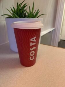 A tall cylindrical red ceramic cup with a white rubber lid that has Costa engraved in bold white writing on it, on a white background.