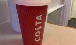 A tall cylindrical red ceramic cup with a white rubber lid that has Costa engraved in bold white writing on it, on a white background.