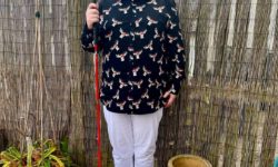 A tall bearded man wearing a black shirt that has a white and red moose print all over it, a pair of white chino trousers and a pair of shiny red shoes holding a bright white cane stood against a dark wooden fence.