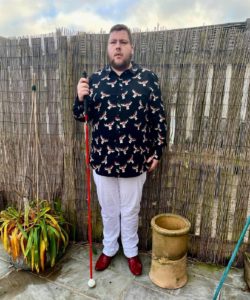 A tall bearded man wearing a black shirt that has a white and red moose print all over it, a pair of white chino trousers and a pair of shiny red shoes holding a bright white cane stood against a dark wooden fence.