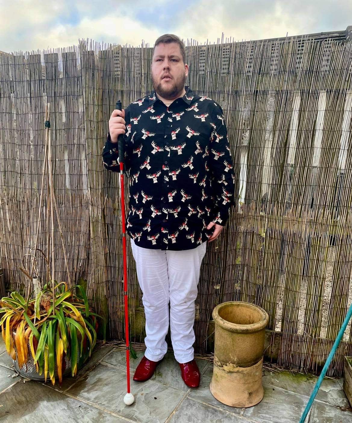 Outfit Of The Day | The Christmas Moose Is On The Loose A tall bearded man wearing a black shirt that has a white and red moose print all over it, a pair of white chino trousers and a pair of shiny red shoes holding a bright white cane stood against a dark wooden fence.
