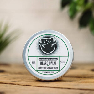 A short cylindrical silver metal tin with a white label that has Beard Monsters Grapefruit & Orange Blast Beard Balm written in bold black writing and a picture of a man with a black beard on it, on a white background.