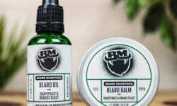 A short cylindrical brown glass bottle with a black rubber pipette lid and a white label that has Beard Monsters Grapefruit & Orange Blast Beard Oil written in bold black writing and a picture of a man with a black beard on it next to a short cylindrical silver metal tin with a white label that has Beard Monsters Grapefruit & Orange Blast Beard Balm written in bold black writing and a picture of a man with a black beard on it, on a white background.