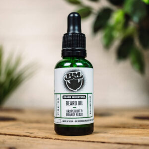 A short cylindrical brown glass bottle with a black rubber pipette lid and a white label that has Beard Monsters Grapefruit & Orange Blast Beard Oil written in bold black writing and a picture of a man with a black beard on it, on a white background.