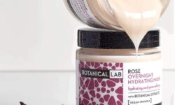 A short cylindrical clear glass jar full of some creamy pink liquid with a black plastic lid and a white label that has a picture of a black stripe, Botanical Lab Rose Overnight Hydrating Mask written in bold black writing, and a picture of some bright pink rose petals on it, on a white background.