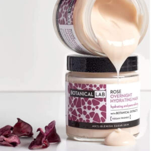 A short cylindrical clear glass jar full of some creamy pink liquid with a black plastic lid and a white label that has a picture of a black stripe, Botanical Lab Rose Overnight Hydrating Mask written in bold black writing, and a picture of some bright pink rose petals on it, on a white background.
