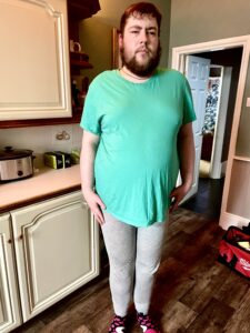 A tall bearded man wearing a bright green crew neck t-shirt, a pair of dark grey leggings and a pair of black and red spotted socks stood against a light grey wall.