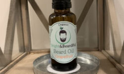 A short cylindrical dark amber glass bottle with a black rubber pipette lid and a white label that has Bright & Beardiful Grapefruit Beard Oil written in bold black writing and a picture of a person with a black beard on it, on a white background.