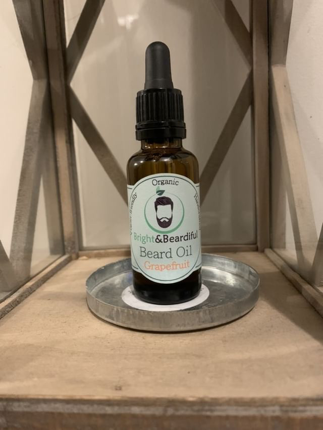 The Bright & Zesty Scent Of Grapefruit | Bright & Beardiful Grapefruit Beard Oil & Beard Butter Combo A short cylindrical dark amber glass bottle with a black rubber pipette lid and a white label that has Bright & Beardiful Grapefruit Beard Oil written in bold black writing and a picture of a person with a black beard on it, on a white background.