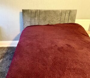 A large rectangular double bed with a rectangular dark grey headboard that's covered in a fluffy burgundy duvet cover, a dark burgundy sheet and two fluffy burgundy pillows, on a white background.