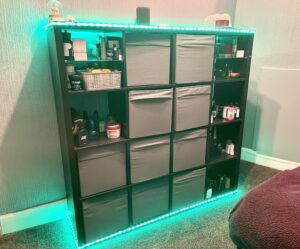 A large rectangular light wooden shelving cabinet full of several large dark grey fabric storage boxes, two clear glass shelves and various different shaped and sized bottles wrapped in some light green coloured strip lights that has a small white polar bear figurine, a large circular snow globe that has a polar bear inside it, a large cylindrical dark grey amazon echo speaker, a small flat google home speaker, a large white polar bear figurine, a small cylindrical grooved glass bowl and a large square white plate on top of it, on a white background.