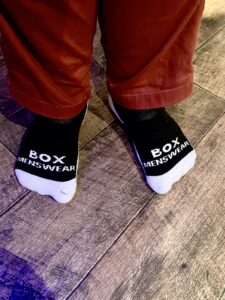 Me wearing a pair of fluffy black socks that has Box Menswear written in bold white writing on them stood on a light wooden laminate floor.