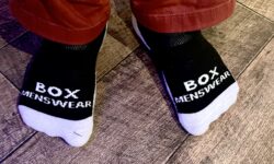 Me wearing a pair of fluffy black socks that has Box Menswear written in bold white writing on them stood on a light wooden laminate floor.