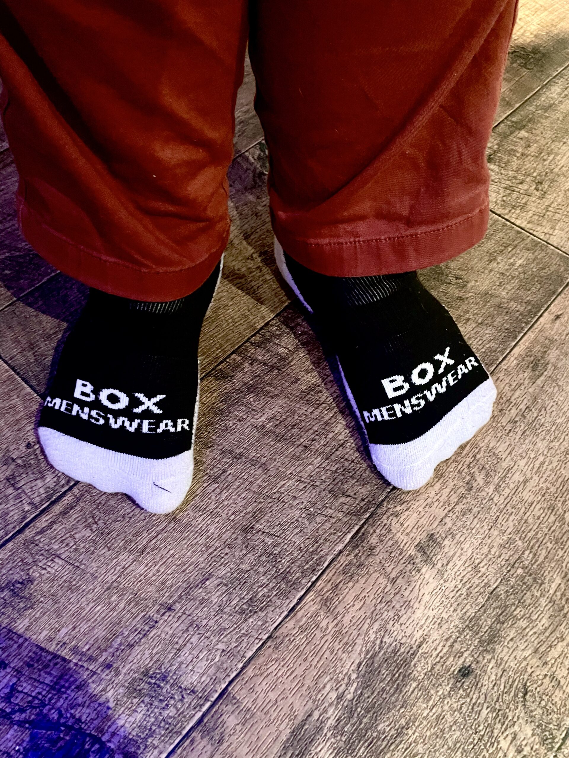 Stand On The Box, Stomp Your Feet | Box Menswear Black Sports Socks Me wearing a pair of fluffy black socks that has Box Menswear written in bold white writing on them stood on a light wooden laminate floor.