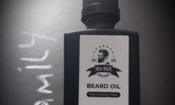 A short rectangular glass Bottle with a black rubber pipette lid and a light brown label that has Man Made Beard Company The Family Man Beard Oil written in bold black writing and a picture of a man with a black beard on it, on a white background.
